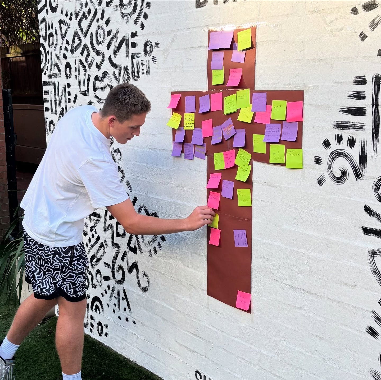 Veta Morphus student adding a sticky note to a large cross on the wall. It is covered in coloured sticky notes. Graffiti art in black and white is behind the cross.